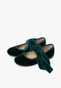 Un par de bailarinas de terciopelo verde oscuro con grandes lazos de terciopelo en la parte superior, plantillas beige y suelas negras sobre un fondo blanco.