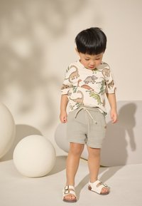 Maglietta chiara con stampe di dinosauri e dettagli fogliari, abbinata a pantaloncini grigi testurizzati e sandali bianchi con doppie fibbie.