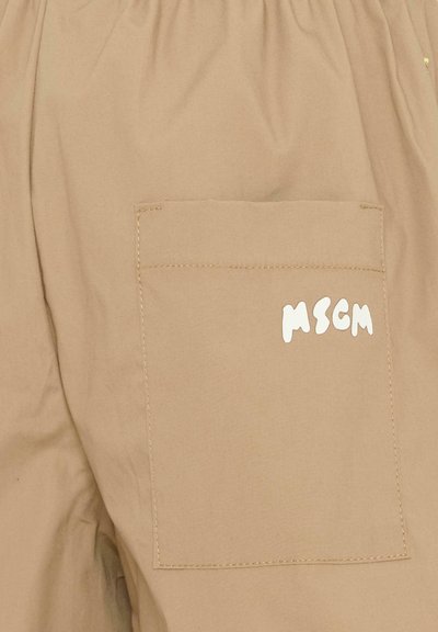 Tissu beige avec une poche carrée cousue présentant des lettres blanches stylisées "MSGM" imprimées près du coin supérieur droit.