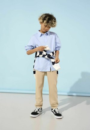 Lichtblauwe button-up shirt, beige broek, zwart-wit gestreepte trui om de taille geknoopt, zwarte sneakers met witte accenten.