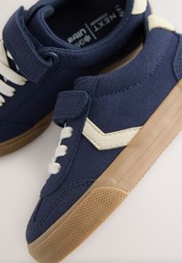 Baskets en daim bleu marine avec accents crème, dotées d'une bride velcro et de lacets blancs. Semelle en gomme texturée et détail de design angulaire.