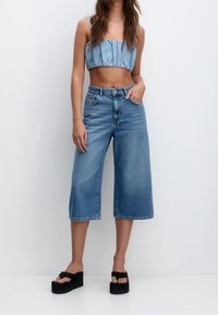 Top corto de denim con detalle fruncido, combinado con culottes azules de gran tamaño. Sandalias de plataforma negras completan el conjunto. Textura suave, estilo casual.