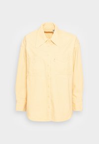 Chemise jaune pâle à manches longues avec col large, boutonnage avant et deux poches poitrine sur fond blanc.