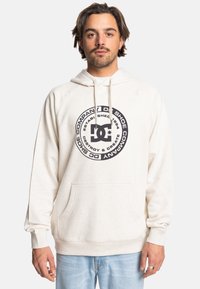 Biała bluza z kapturem z okrągłym czarnym logo, kieszenią z przodu i sznurkiem do regulacji. Materiał wydaje się być miękki. Model ma na sobie jasne dżinsy.