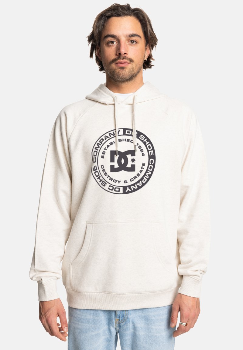 Biała bluza z kapturem z okrągłym czarnym logo, kieszenią z przodu i sznurkiem do regulacji. Materiał wydaje się być miękki. Model ma na sobie jasne dżinsy.