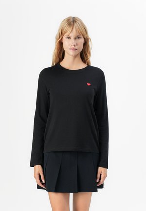 Femme blonde portant un t-shirt noir à manches longues avec un petit cœur rouge sur la poitrine et une jupe plissée noire, debout sur fond blanc.