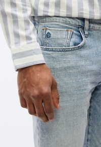 Κοντινό πλάνο ενός χεριού που ξεκουράζεται κοντά στην μπροστινή τσέπη ενός ανοιχτόχρωμου τζιν παντελονιού, με ορατή τη μανσέτα πουκαμίσου με ρίγες.