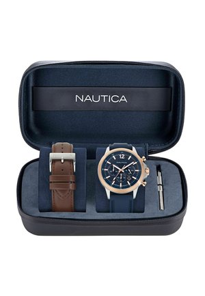 Montre Nautica dans un étui zippé noir. Comprend une montre en silicone navy avec des accents en or rose et un bracelet en cuir marron. Inclus un outil d'ajustement.