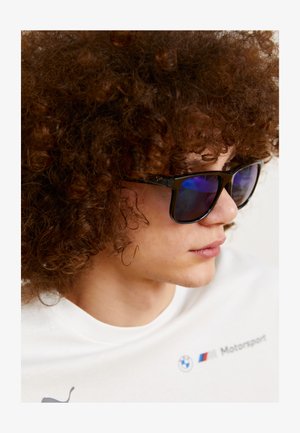 Lunettes de soleil noires avec des verres teintés bleus, dotées d'une monture carrée. Le modèle a des cheveux bouclés bruns et porte un t-shirt blanc avec des détails de logo.