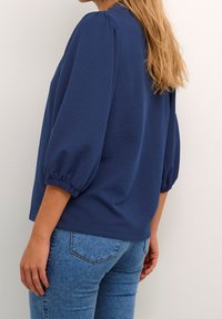 Blusa over-size blu navy in tessuto testurizzato, con maniche a palloncino arricciate e scollatura rotonda, abbinata a jeans aderenti blu chiaro.