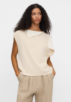 Kobieta o kręconych włosach ubrana w beżową bezrękawnik z asymetrycznym kołnierzem oraz beżowe plisowane spodnie, ręce w kieszeniach, neutralne tło.