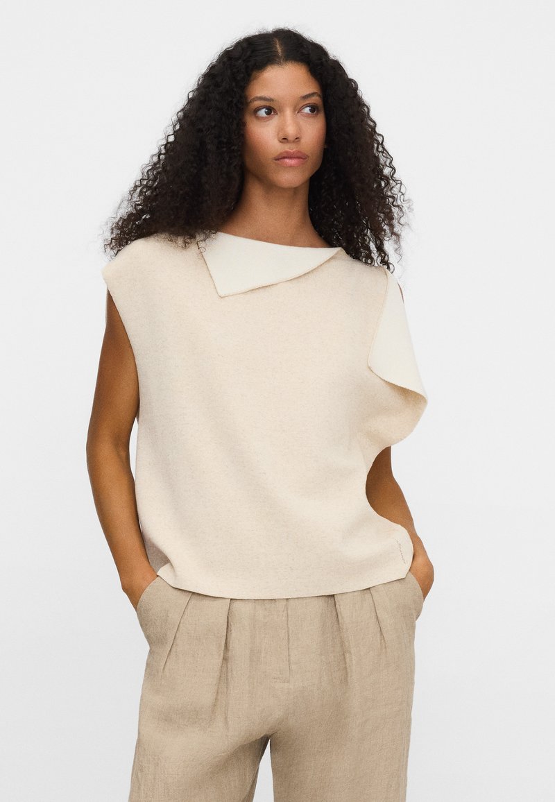 Kobieta o kręconych włosach ubrana w beżową bezrękawnik z asymetrycznym kołnierzem oraz beżowe plisowane spodnie, ręce w kieszeniach, neutralne tło.