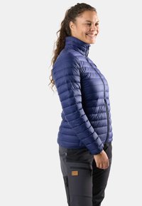 Tummansininen untuvatakki, jossa on korkea kaulus, vaakasuorat tikkaukset ja vetoketju. Yhdistetty tummiin housuihin ja logopatchilla varustettuun taskuun.