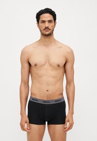 Schwarze Boxershorts aus elastischem Stoff mit einem grauen Gummibund, der das Diesel-Logo trägt. Das Modell steht mit einem neutralen Gesichtsausdruck.