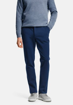 Mann in navyblauen Hosen, hellgrauen Sneakers und einem blauen Langarmpullover, der mit einer Hand in der Tasche vor einem weißen Hintergrund steht.