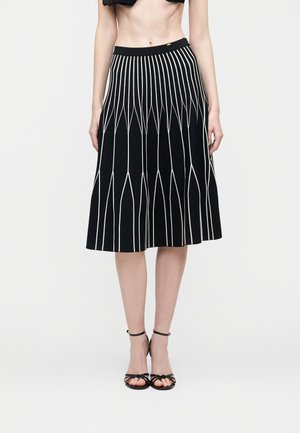 Jupe noire longueur genou avec des lignes géométriques verticales blanches portée avec des sandales à talons hauts noires à brides, le ventre du mannequin visible.