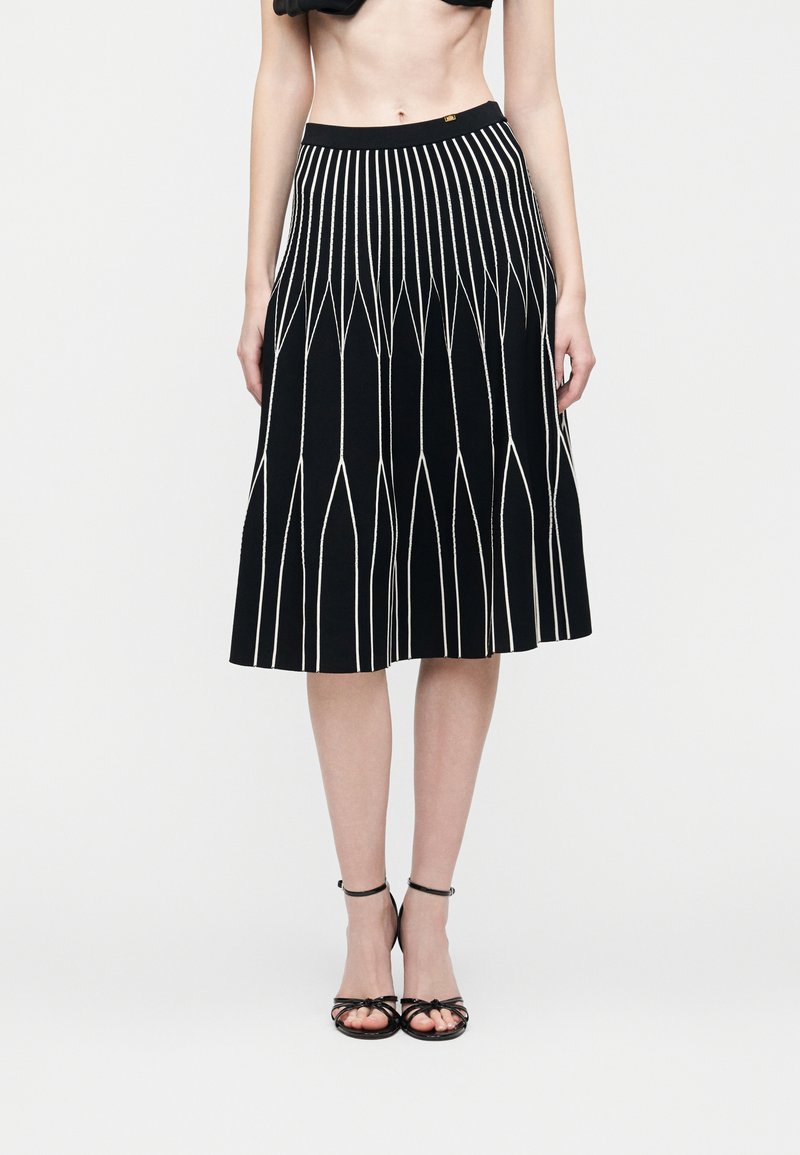 Μαύρη φούστα μέχρι το γόνατο με λευκές γεωμετρικές κάθετες γραμμές, φορεμένη με μαύρα πέδιλα με λουράκια και τακούνι, με εμφανές το μέσο της μέσης του μοντέλου.