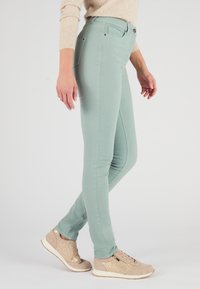 Vaqueros skiny verde claros con una textura suave y diseño de cinco bolsillos, combinados con zapatillas beige que tienen un acento metálico.