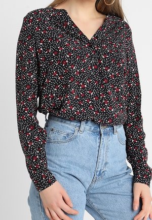 Vrouw draagt een zwarte blouse met lange mouwen, rood-wit bloemenpatroon, weggestopt in lichtblauwe spijkerbroek met hoge taille.