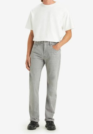 Person trägt ein weißes übergroßes T-Shirt, hellgraue Jeans mit geradem Schnitt, schwarze Turnschuhe, steht mit einer Hand in der Tasche.