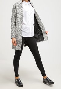 Abrigo largo de frente abierto en un patrón salpicado de blanco y negro, que se superpone a una camisa blanca y unos jeans skinny negros, finalizado con zapatos de tacón negros.