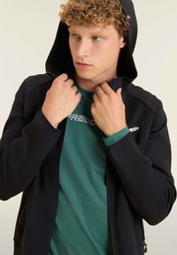 Sudadera negra con cremallera y capucha, llevada sobre una camiseta verde con un logotipo. Se pueden ver el material texturizado y los puños verdes en contraste.