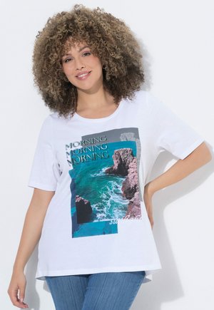 Frau mit lockigem Haar lächelnd, trägt weißes T-Shirt mit Ozean- und Klippengrafik und blaue Jeans, steht vor weißem Hintergrund.