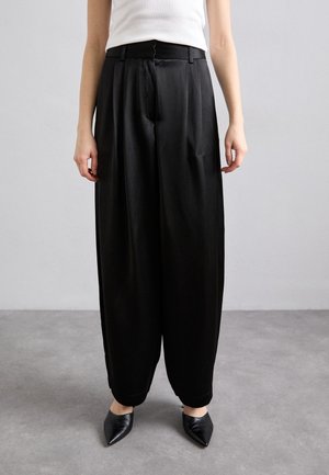 Mujer con pantalones negros de cintura alta y pernera ancha con cremallera frontal, combinados con una camiseta blanca sin mangas y zapatos negros puntiagudos.