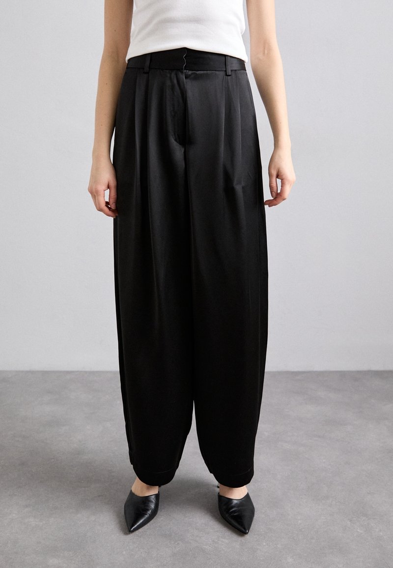 Femme portant un pantalon noir taille haute à jambes larges avec une fermeture éclair devant, associé à un haut blanc sans manches et des chaussures pointues noires.