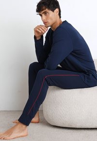 Marineblaues Langarmoberteil und passende Hose im Slim-Fit mit roten Seitenstreifen. Weicher Stoff, lässiger Schnitt, barfuß getragen.