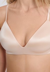 Bralette beige doux avec une finition satinée lisse, des bonnets triangulaires, des bretelles ajustables et un design en V, mettant en avant des coutures minimalistes.
