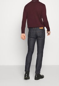 Mörkblå denimjeans med en figursydd design, bakficka märkt "DIESEL" och kontrastsömmar. Afrikad med en vinröd långärmad skjorta.