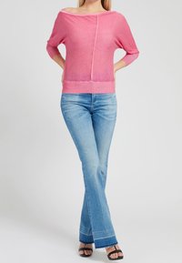 Suéter rosa claro sin mangas con mangas de tres cuartos, combinado con jeans azules acampanados. La vestimenta presenta un corte relajado y una textura suave.