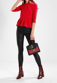 Kobieta w czerwonej bluzce peplum, czarnych dopasowanych spodniach, czerwonych botkach za kostkę, trzymająca czarną i czerwoną torbę z złotym zapięciem, stojąca na białym tle.
