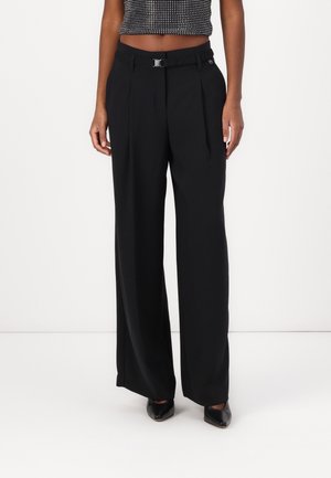 Woman wearing high-waist black wide-leg trousers with pleats and a black belt, paired with black pointed-toe shoes and a cropped top.