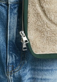 Beige fleece vest met groene afwerking, met een zilveren rits. Het vest is gelaagd over blauwe denim met zichtbare stiksels.