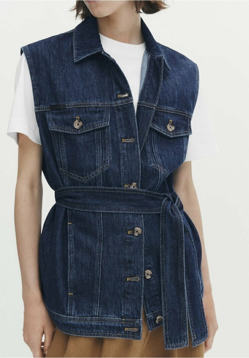 Femme portant un gilet en jean bleu foncé sans manches avec des boutons devant, des poches poitrine, une ceinture nouée à la taille, par-dessus une chemise blanche à manches courtes.