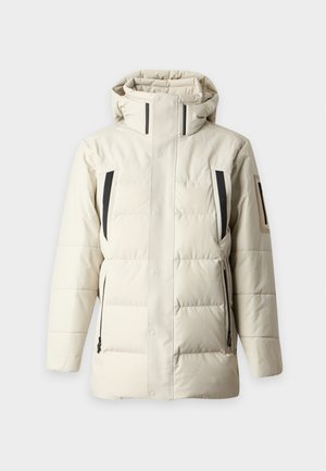 Homme portant une doudoune beige clair par-dessus un pull bleu à fermeture éclair et un pantalon noir, debout les mains dans les poches devant un fond uni.