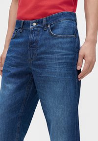 Homme portant un jean en denim bleu et une chemise rouge, les mains détendues le long du corps devant un fond clair uni.