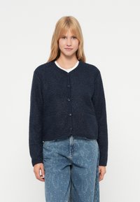 Marineblå, teksturert cardigan med rund hals, knapping foran og to sidelommer, brukt over en hvit t-skjorte.