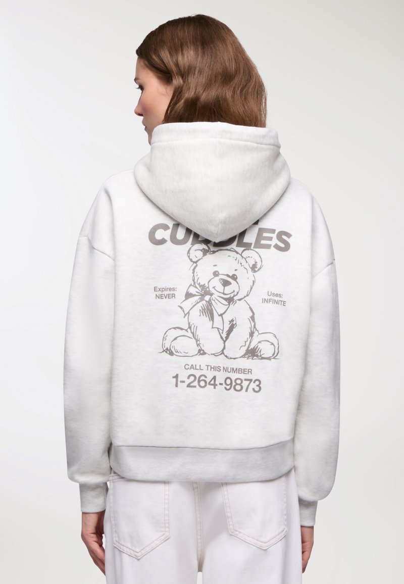 Donna con capelli castani che indossa un felpa bianco con un disegno di un orsetto e la scritta "CUDDLES" e un numero di telefono sul retro.