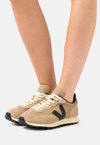 Beige semskinn sneakers med svarte detaljer, rund tå og kremfargede snører. Teksturert sidepaneler og en robust svart såle.