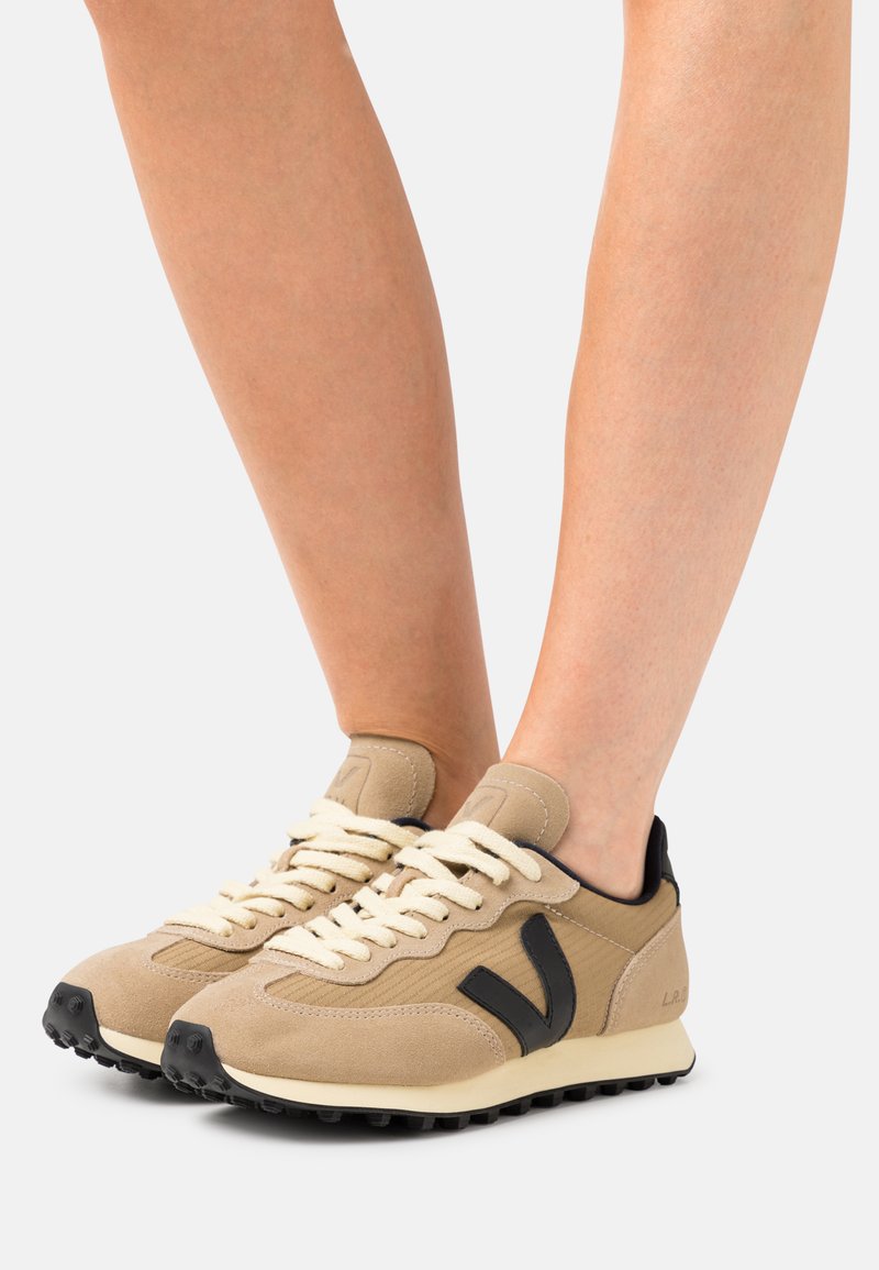 Beige semskinn sneakers med svarte detaljer, rund tå og kremfargede snører. Teksturert sidepaneler og en robust svart såle.