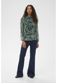 Göndör hajú nő zöld mintás fodros blúzban, sötét szárszélesített farmerral és fekete cipőben, egyszerű háttér előtt állva.