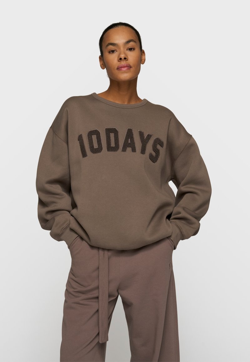 Femme debout, les mains dans les poches, portant un sweat-shirt marron oversize avec le texte « 10DAYS » et un pantalon assorti marron, devant un fond uni.