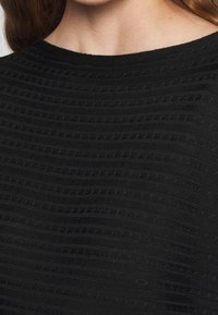 Svart topp med en texturerad, ribbad design med horisontella linjer och små öppningar, som har en rund halslinje och mjukt stickat material.