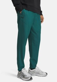 Atletske hlače v barvi zelene modrine s slim krojem, ob strani imajo žepke in diskretno črno logotip. Material je gladek in diha, z lastnostmi za vpijanje vlage.