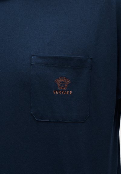 Tmavě modrá látka s náprsním kapesníkem vyšívaným logem hlavy Medúzy od Versace a názvem značky v hnědém vlákně.