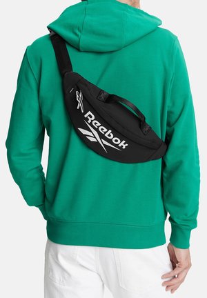 Persona che indossa una felpa verde con cappuccio e pantaloni bianchi, con una borsa a tracolla nera Reebok indossata sulla schiena.