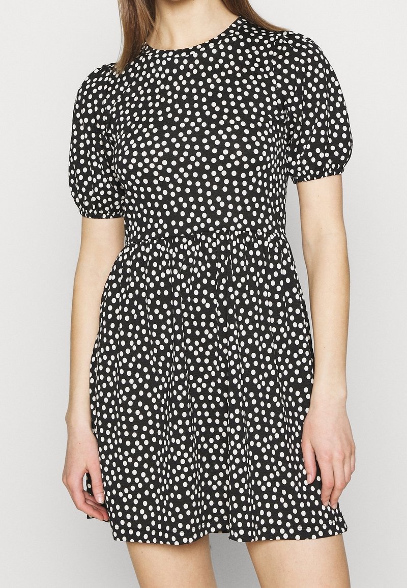 Vestido negro con lunares blancos, mangas cortas abullonadas, escote redondo y cintura fruncida. Hecho de tela ligera, hasta la rodilla.